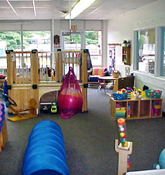 A child care room at the center filled with toys, a wooden play structure and lots of natural light coming from a wall of windows at the back of the room