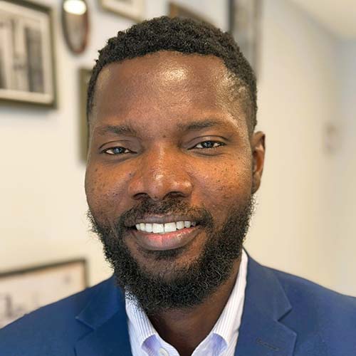 Man with a blue suit jacket standing inside in front of a gallery wall smiles brightly for the camera.