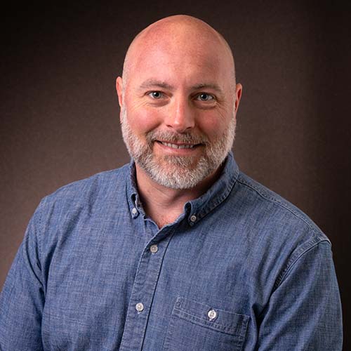 Man with a blue denim shirt sits for a professional headshot, smiling brightly
