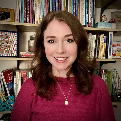 Woman with a bright, deep fuchsia sweater sits for a photograph in front of a large, full bookshelf.