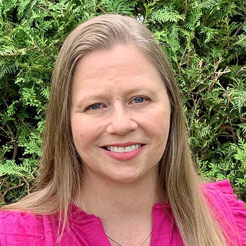 Woman with fuchsia pink shirt stands outside in front of a green hedge for a professional headshot