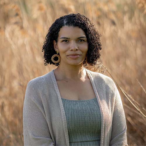 Woman with chin-length dark, curly hair stands in earth-toned clothes in front of a field of brown grasses