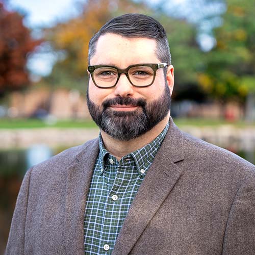 Man with dark brown glasses and brown sports coat stands outside in front of an academic building. Blue sky peppered with white clouds in the background.