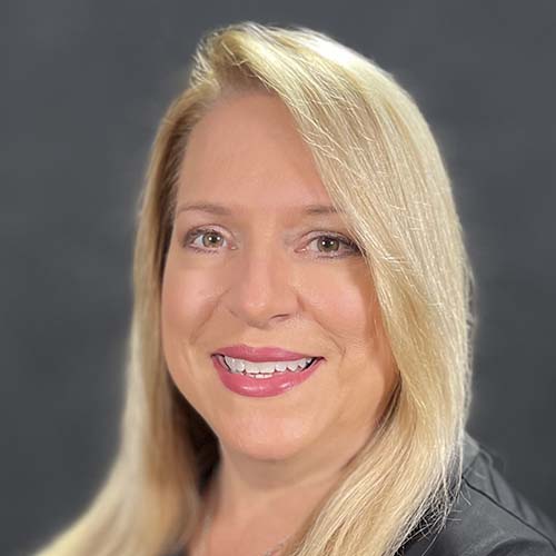 Woman with long blond hair in a dark dress shot against a dark background. She smiles for the camera