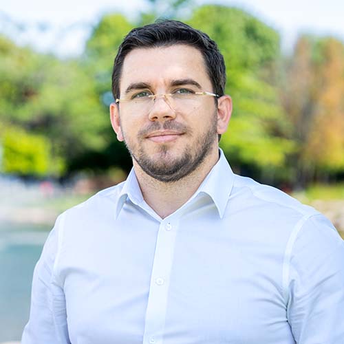 Man in a light blue button-up shirt stands outside in front of a pond for a headshot.