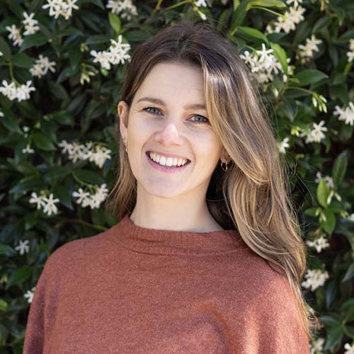 Woman stands outside against a flowering hedge, smiling for the camera