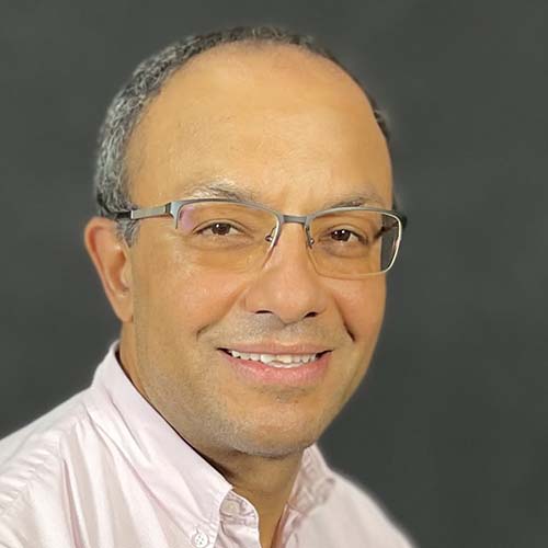 Man with a light pink button-up shirt sits for a headshot against a gray background.