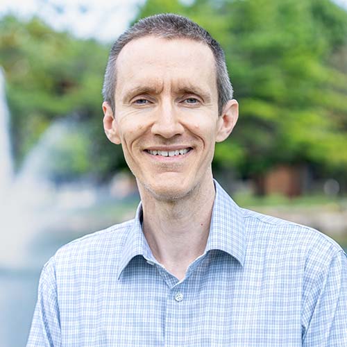 Man in a blue plaid button-up shirt stands outside for a professional headshot