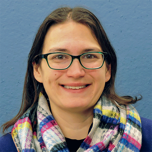 Woman with long dark hair and blue rimmed glasses stands against a sky blue background wearing a colorful scarf.