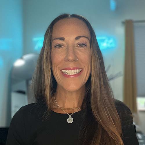 Woman with long dark hair in a studio with a blue neon sign illuminated behind her. She smiles brightly.