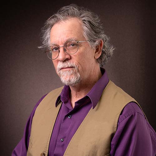 Man with a jewel-tone purple shirt and brown vest sits stoicly for a photo