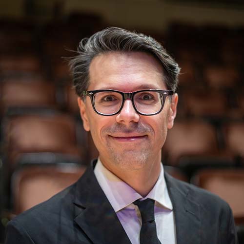 Man standing on a concert stage with a suit and tie, smiles without teeth for the camera.