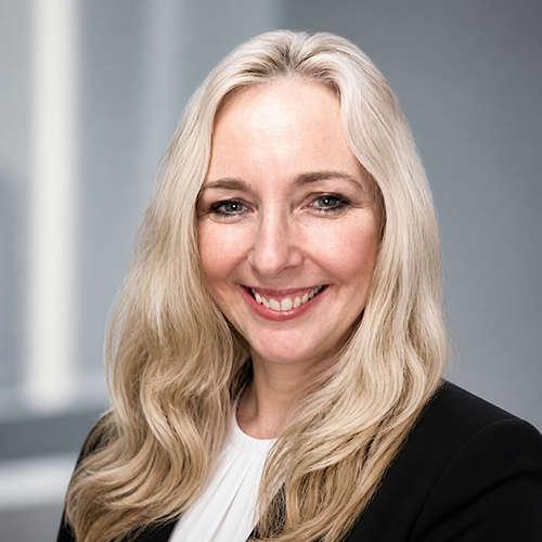Woman with long, light hair wears a black blazer and sits for a professional headshot.