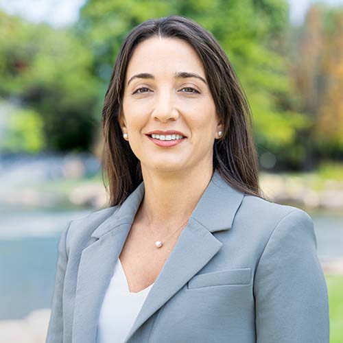 Woman with long dark hear wearing a gray suit jacket stands outside for a professional headshot