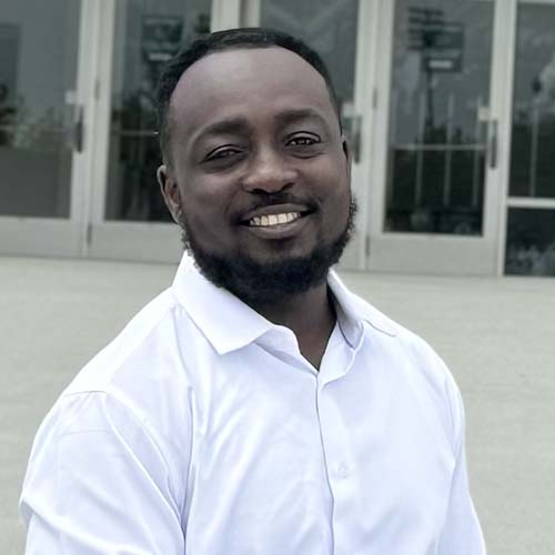 Man standing outside in front of an academic building, smiles brightly for the camera.