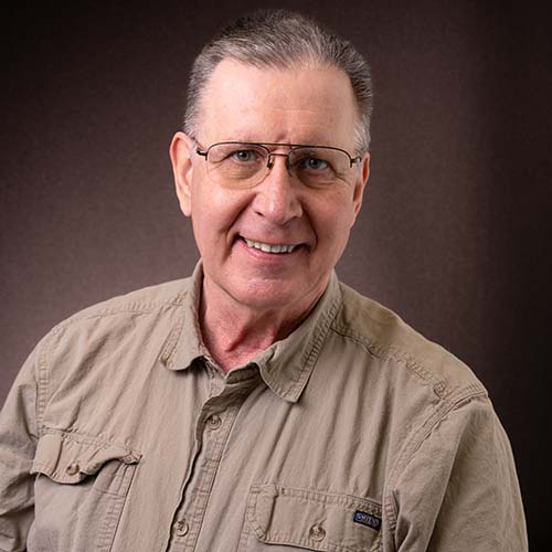 Man with a earthy-brown shirt and yellow-tinted glasses sits for a professional headshot