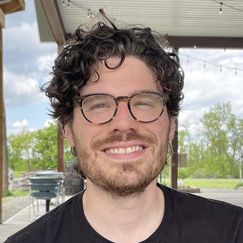 Man with curly dark hair wearing round tortoise shell glasses sits outside, smiling for picture.