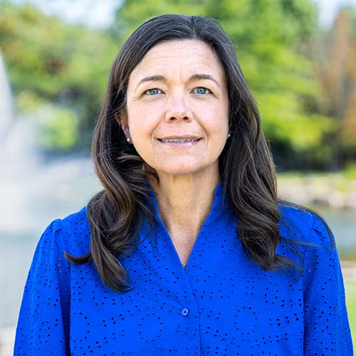 Woman with long dark hair and a bright blue eyelet blouse stands outside by Hawkins Pond for a professional headshot.