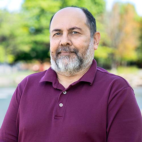 Man with a dark beard sits outside by Hawkins Pond for a professional headshot.