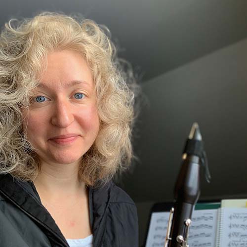 A woman with curly shoulder length blond hair and light blue eyes is on the left of the frame, holding her clarinet.