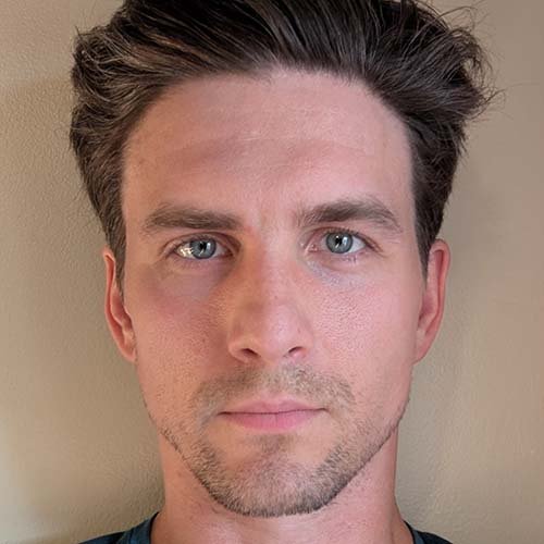 Close-up headshot of a man with blue eyes and stubble against a wall.