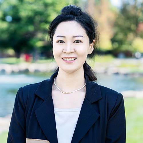Woman with dark hair pulled back wearing a dark suit jacket smiles brightly for a headshot in front of Hawkins Pond.