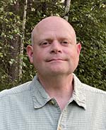 man with light green collared shirt stands in front of trees
