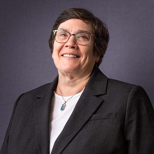 Woman with short dark hair and glasses wearing a professional suit coat smiles brightly for the camera