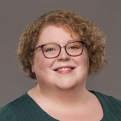 Woman with chin-length curly red hair sits for a professional headshot. She has a friendly smile.