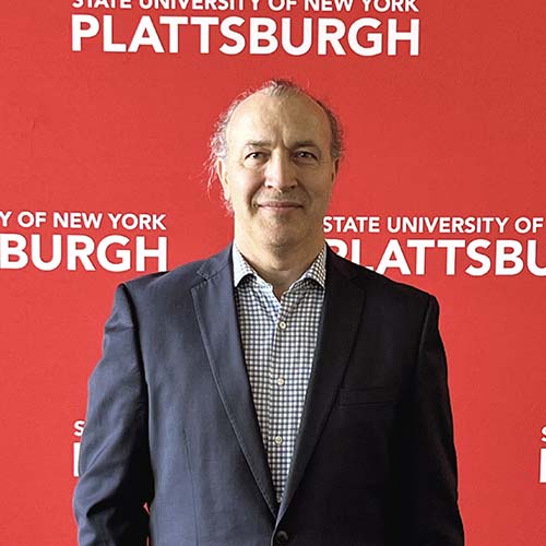 Man in a dark suit standing in front of a SUNY Plattsburgh photo background
