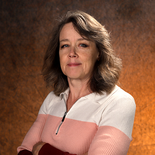 Woman stands in a dimly lit studio, posing with a slight sideways turn, arms folded.