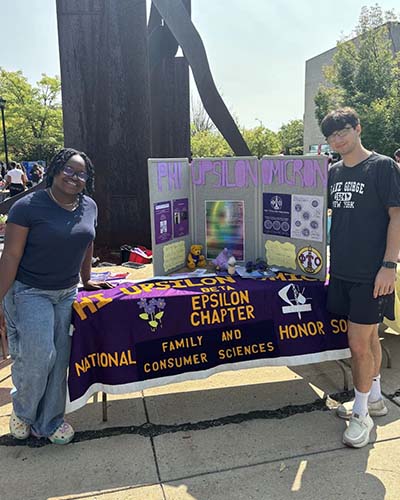 Two students stand outside during the activities fair with Phi Upsilon Omicron materials at a table.