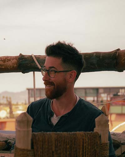 Man with glasses and a beard smiling while sitting outdoors at dusk; a woven natural-fiber chair is visible in the foreground and buildings appear blurred in the background.